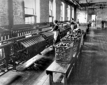 Three women in long black dresses with white aprons stand in front of a large contraption known as a spinning mule. The spinning mule made cotton into yarn. Baskets of cotton yarn are on benches behind the women.