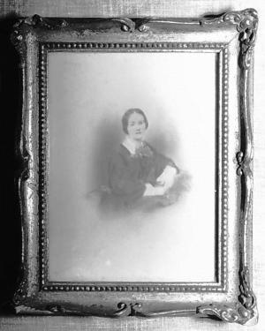 A woman in a dark colored dress is sitting looking toward the viewer.  She is wearing a dark dress with white at the collar and the wrists.  Her dark hair is parted in the middle and pulled back and secured at her nape.